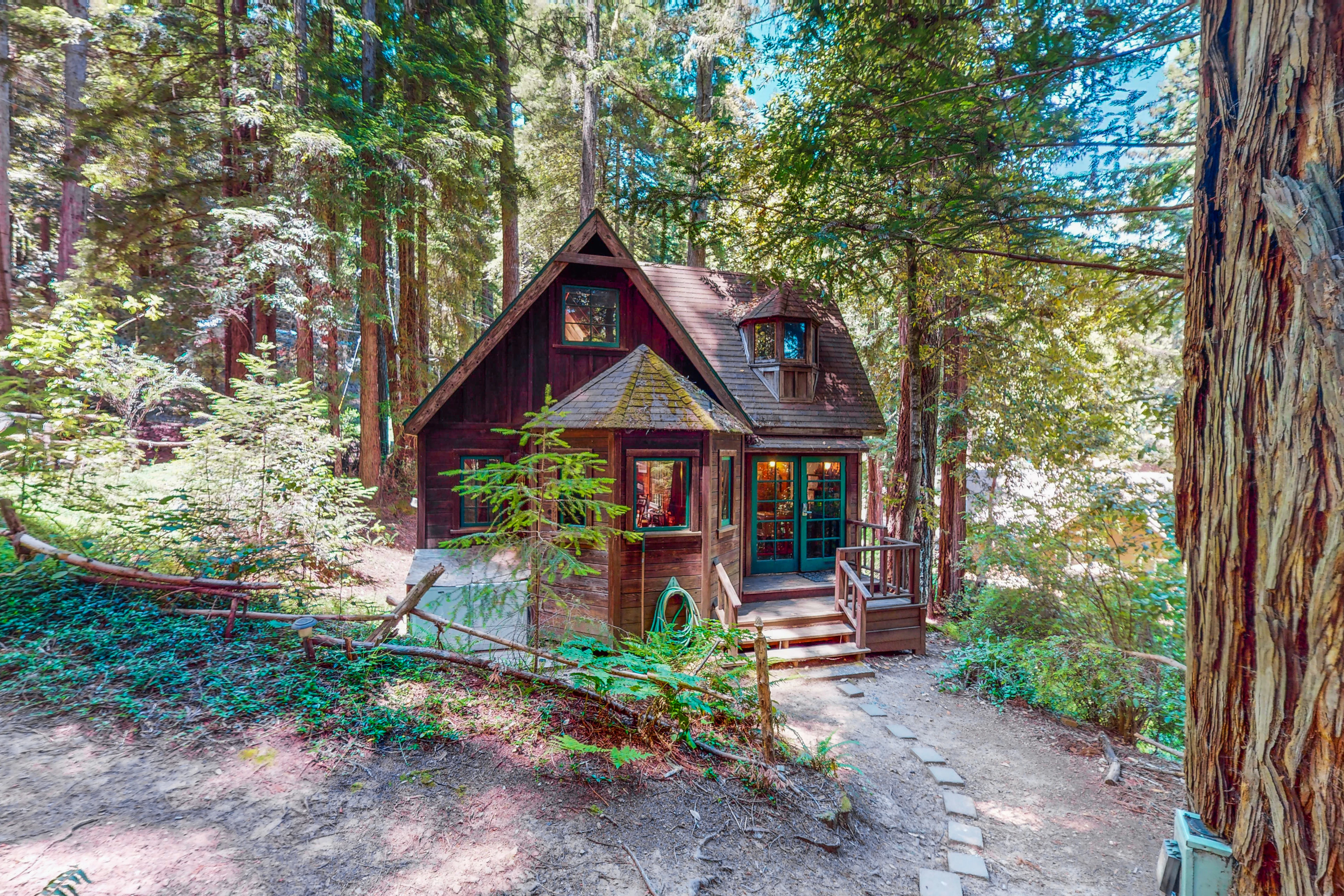 Tucked beneath towering Cazadero redwoods, your storybook cottage awaits — whimsical A-frame charm, warm wood siding, and a stone pathway leading to your private forest hideaway.