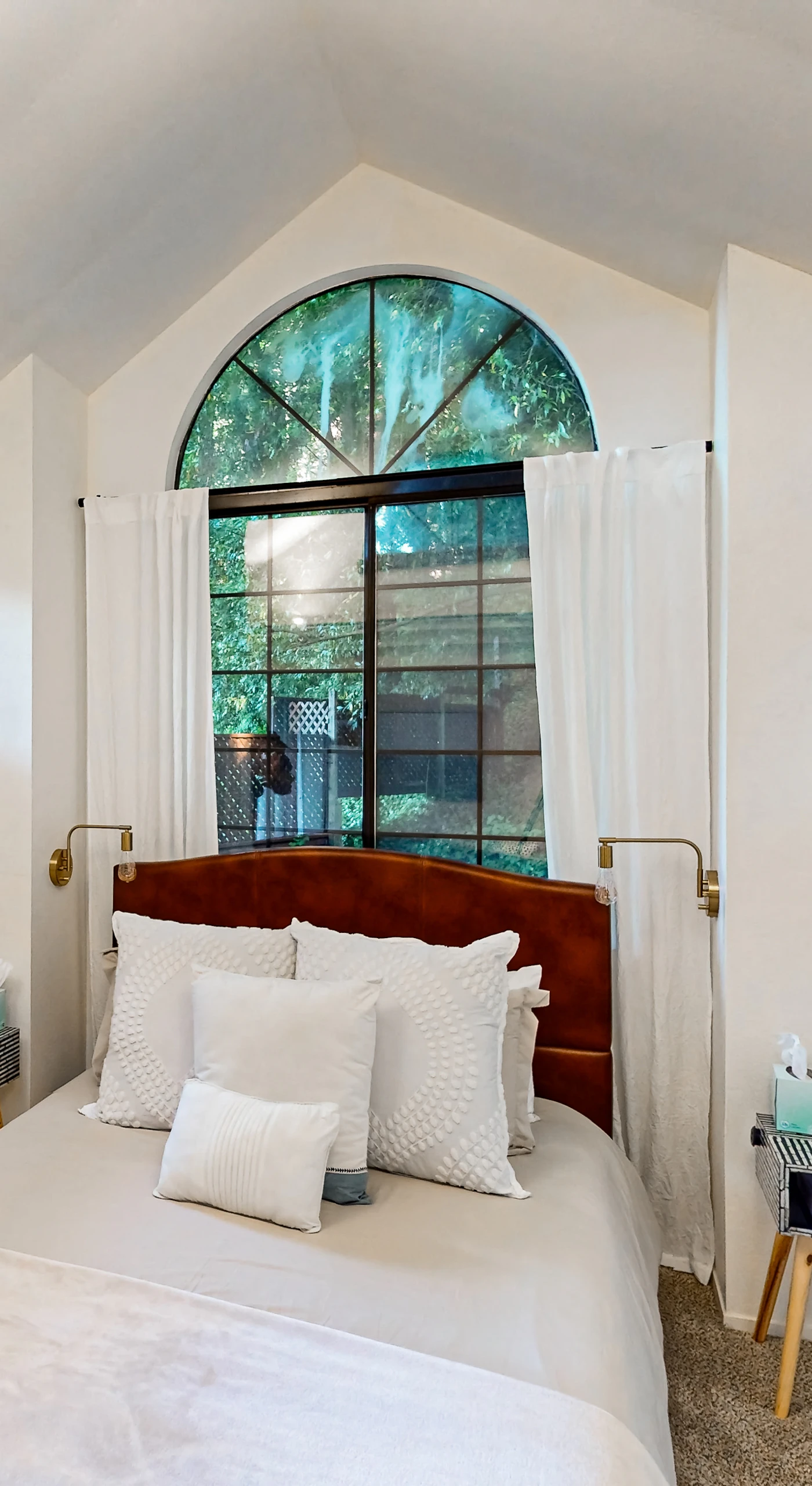 Rest easy in your serene bedroom — a queen bed with crisp white linens sits beneath a stunning arched window that frames the redwood canopy above Monte Rio.