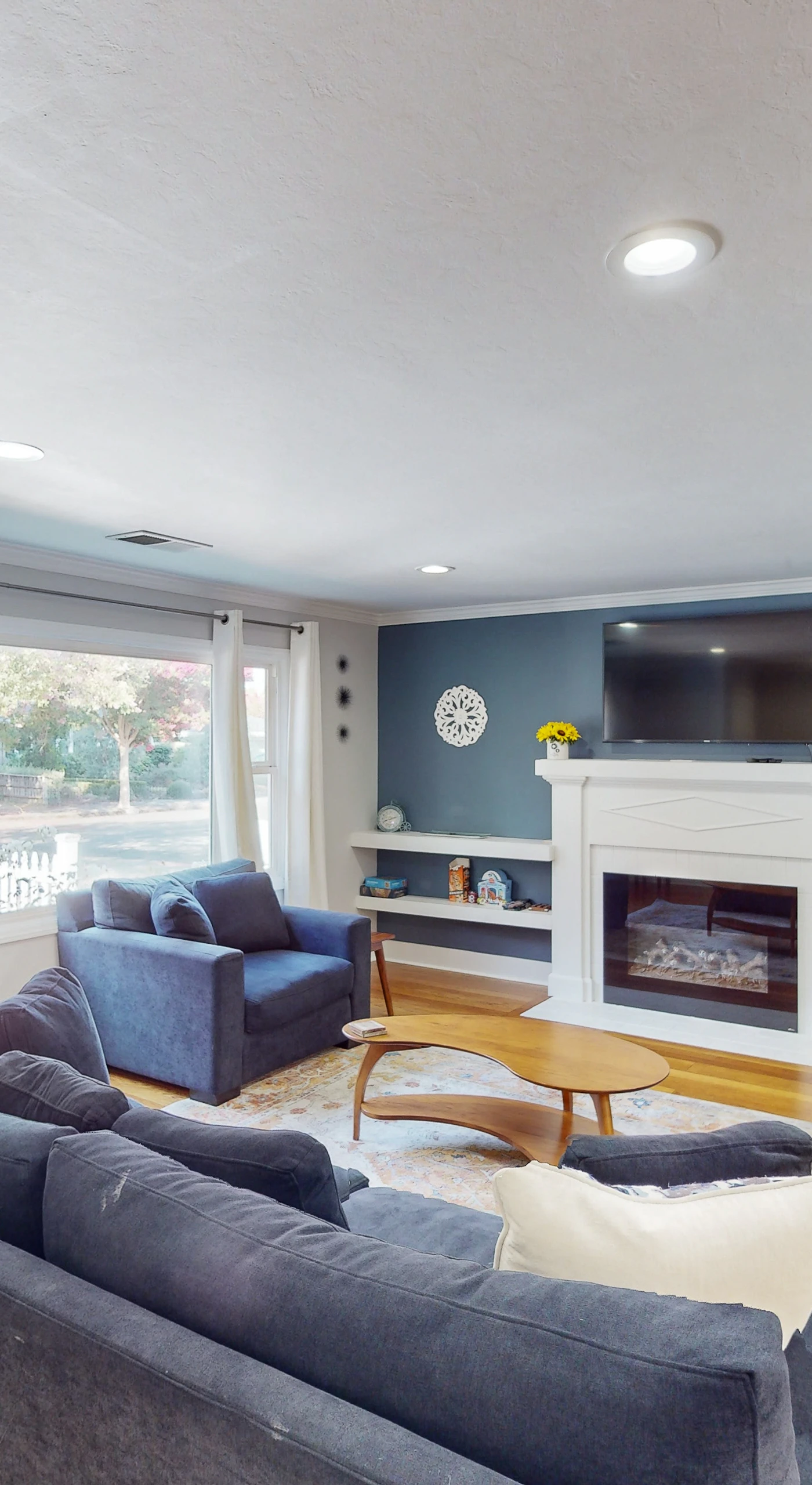 Gather around the fireplace in a sunlit living room with a bold blue accent wall, wall-mounted TV, built-in shelving, and plenty of plush seating for your group
